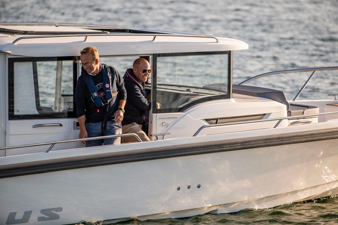 Two men on a modern boat, one standing and laughing while the other sits at the helm, with water in the background.